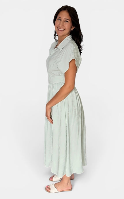 A woman modeling a light sage green midi dress with short sleeves, a collared neckline, and a pleated skirt, standing against a plain white studio background.