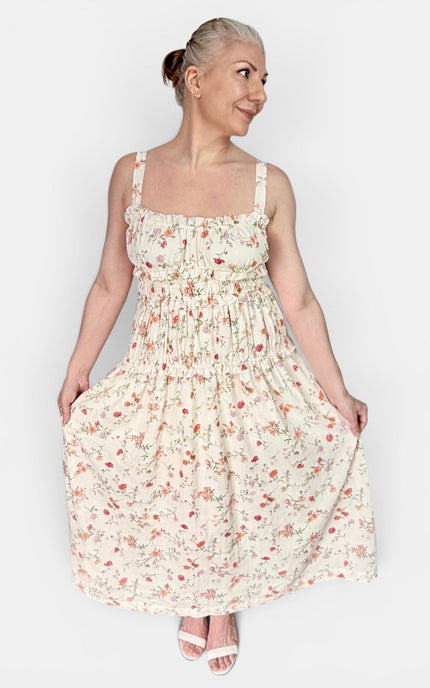 A woman modeling a sleeveless, smocked, tiered maxi dress with a small floral print on a cream background, paired with white heeled sandals, against a plain light gray studio background.