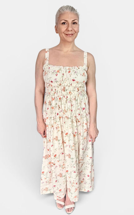 A woman modeling a sleeveless, smocked maxi dress with a delicate pink and red floral print on a cream background, set against a plain light gray studio background.