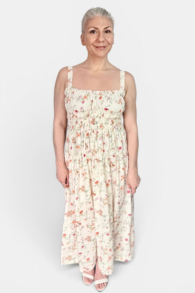 A woman modeling a sleeveless, smocked maxi dress with a delicate pink and red floral print on a cream background, set against a plain light gray studio background.