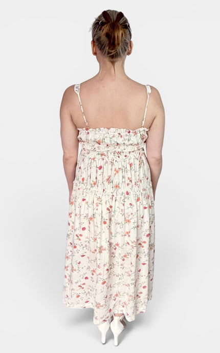 A woman viewed from behind wearing a sleeveless midi dress with thin straps, a smocked bodice, and a small pink floral print on a cream background, paired with white pointed shoes against a light gray studio background.