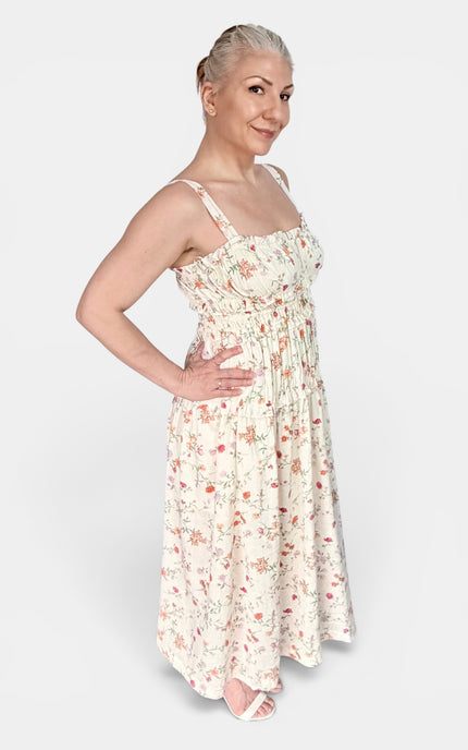 A woman modeling a sleeveless, smocked-waist maxi dress with a delicate red and green floral print on a cream background, set against a plain light gray studio background.