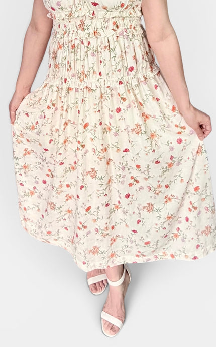A woman wearing a cream-colored tiered maxi dress with a delicate red and green floral print, paired with white heeled sandals, shown against a plain light gray background.