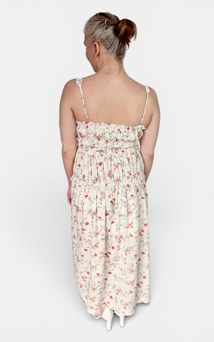 A woman viewed from the back wearing a sleeveless, smocked-waist maxi dress with thin straps and a delicate pink and green floral print on a cream background, set against a plain light gray studio backdrop.