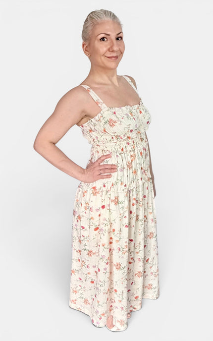 A woman modeling a sleeveless, smocked-bust maxi dress with a delicate pink and red floral print on a cream background, set against a plain light gray studio background.