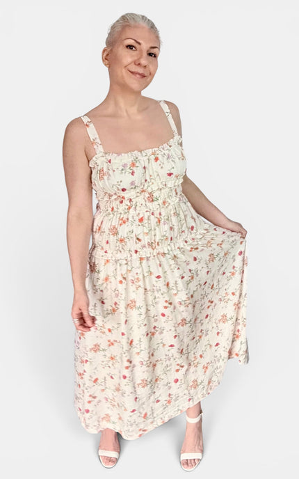 A woman modeling a sleeveless midi dress with thin straps and a small floral print on a light cream background, paired with white heeled sandals, against a plain light gray studio background.