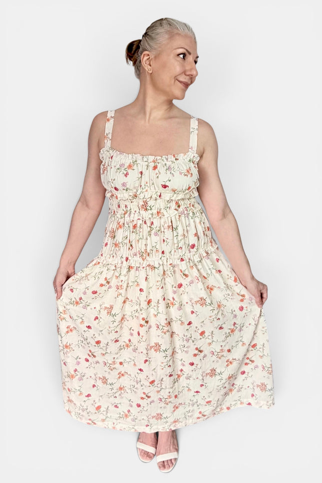A woman modeling a sleeveless, smocked, tiered maxi dress with a small floral print on a cream background, paired with white heeled sandals, against a plain light gray studio background.