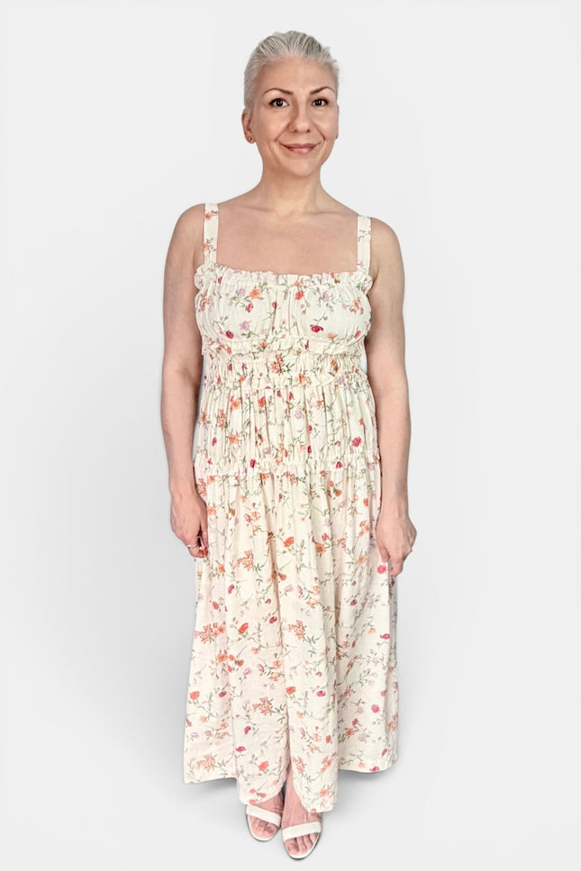 A woman modeling a sleeveless, smocked maxi dress with a delicate pink and red floral print on a cream background, set against a plain light gray studio background.