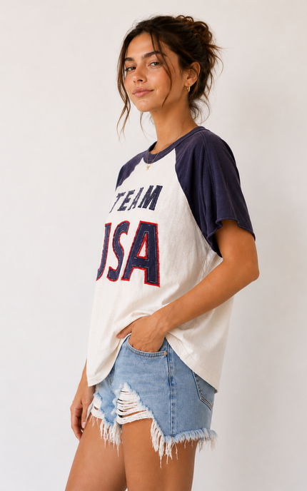 Woman wearing a 'Team USA' baseball-style t-shirt and denim shorts on a plain background
