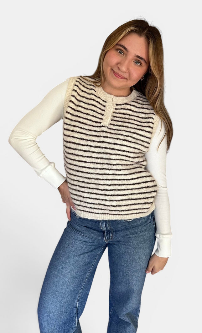 A woman modeling a sleeveless knit vest with horizontal black and cream stripes over a cream long-sleeve shirt and blue jeans, against a light gray studio background.