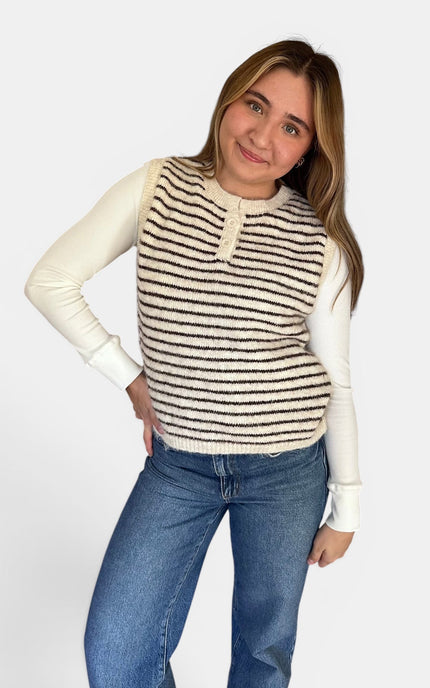 A woman modeling a sleeveless knit vest with horizontal black and cream stripes over a cream long-sleeve shirt and blue jeans, against a light gray studio background.
