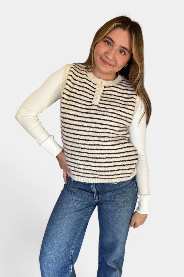 A woman modeling a sleeveless knit vest with horizontal black and cream stripes over a cream long-sleeve shirt and blue jeans, against a light gray studio background.