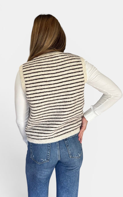 A woman shown from the back wearing a cream knit striped sweater vest over a white long-sleeve shirt and blue jeans, against a plain light gray studio background.