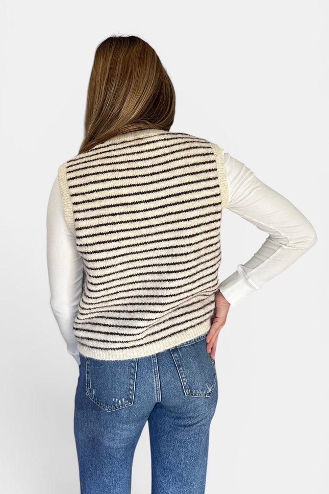 A woman shown from the back wearing a cream knit striped sweater vest over a white long-sleeve shirt and blue jeans, against a plain light gray studio background.