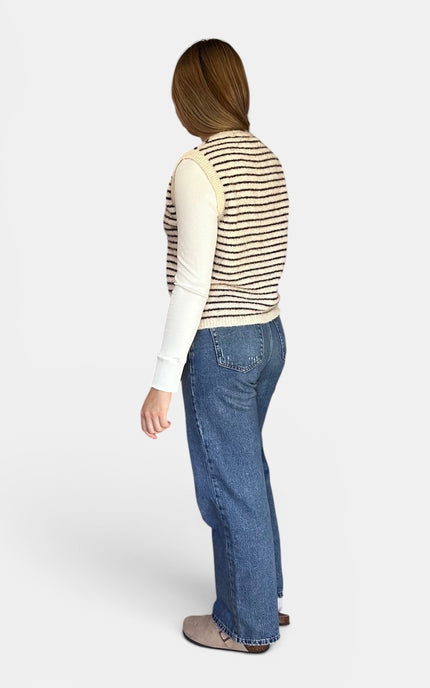 A woman modeling a white long-sleeve top with thin black horizontal stripes and wide-leg blue jeans, shown from a side-back angle against a light gray studio background.