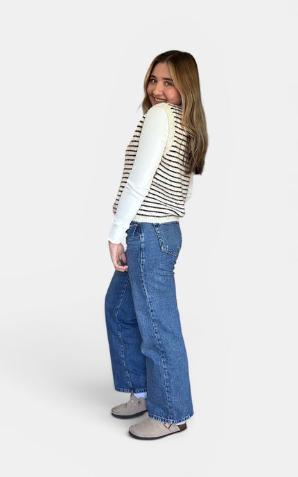 A woman modeling a white long-sleeve top with thin black horizontal stripes and wide-leg blue jeans, shown from a side-back angle against a light gray studio background.