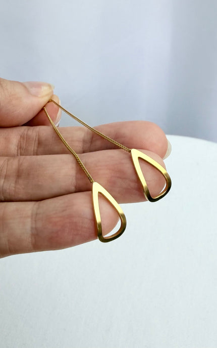 A minimal, handheld shot of a pair of gold geometric threader earrings in a palm. The tight crop focuses on the clean, architectural lines of the open triangle charms and the high-quality, tarnish-free gold finish.