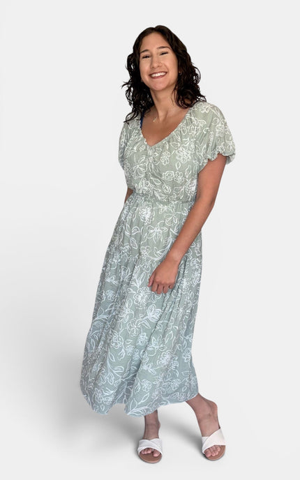 Light sage green and white floral print midi dress with short puff sleeves and a V-neckline, paired with white slide sandals, against a plain light gray studio background.
