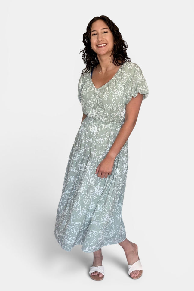 Light sage green and white floral print midi dress with short puff sleeves and a V-neckline, paired with white slide sandals, against a plain light gray studio background.