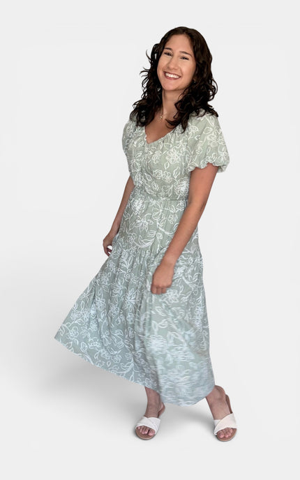 Sage green and white floral print midi dress with short puff sleeves and a V-neckline, paired with white slide sandals, against a plain light gray studio background.