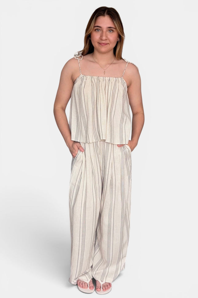 A woman modeling a two-piece matching set consisting of a sleeveless top with thin shoulder straps and wide-leg pants, both in a cream-colored fabric with vertical gray stripes, set against a plain light gray background.