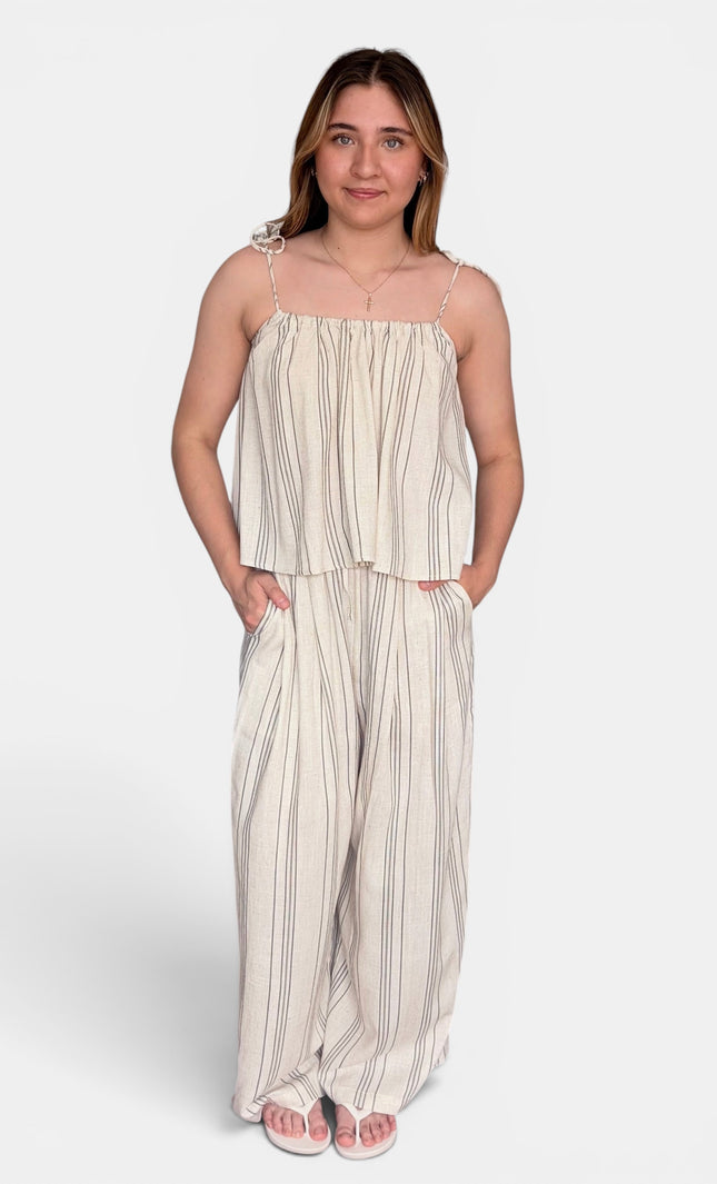 A woman modeling a two-piece matching set consisting of a sleeveless cream and beige vertically striped crop top with spaghetti straps and matching wide-leg pants, set against a plain light gray background.