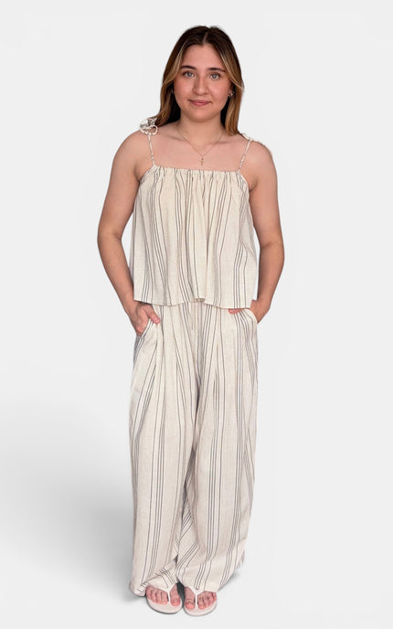 A woman modeling a two-piece matching set consisting of a sleeveless cream and beige vertically striped crop top with spaghetti straps and matching wide-leg pants, set against a plain light gray background.