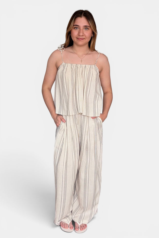 A woman modeling a two-piece matching set consisting of a sleeveless cream and beige vertically striped crop top with spaghetti straps and matching wide-leg pants, set against a plain light gray background.