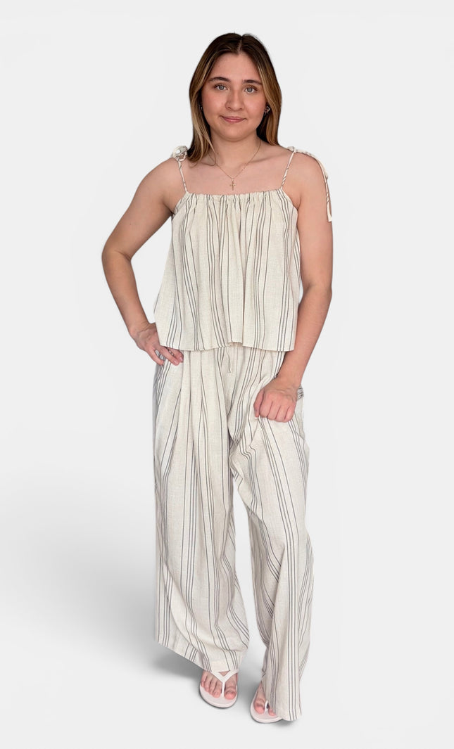 A woman modeling a two-piece matching set consisting of a sleeveless cream and beige vertically striped peplum top and wide-leg trousers, styled with white sandals against a light gray studio background.