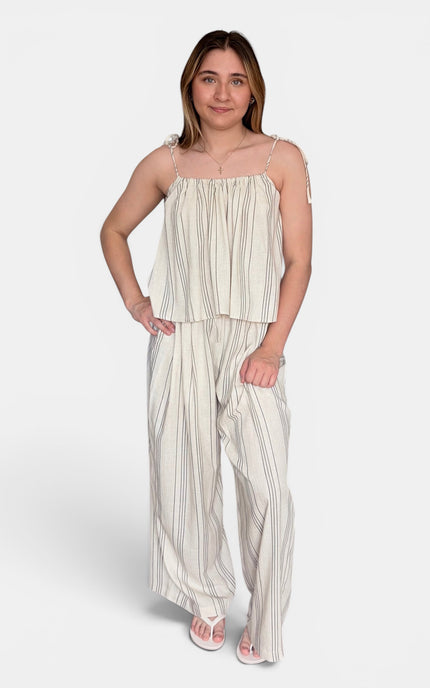 A woman modeling a two-piece matching set consisting of a sleeveless cream and beige vertically striped peplum top and wide-leg trousers, styled with white sandals against a light gray studio background.