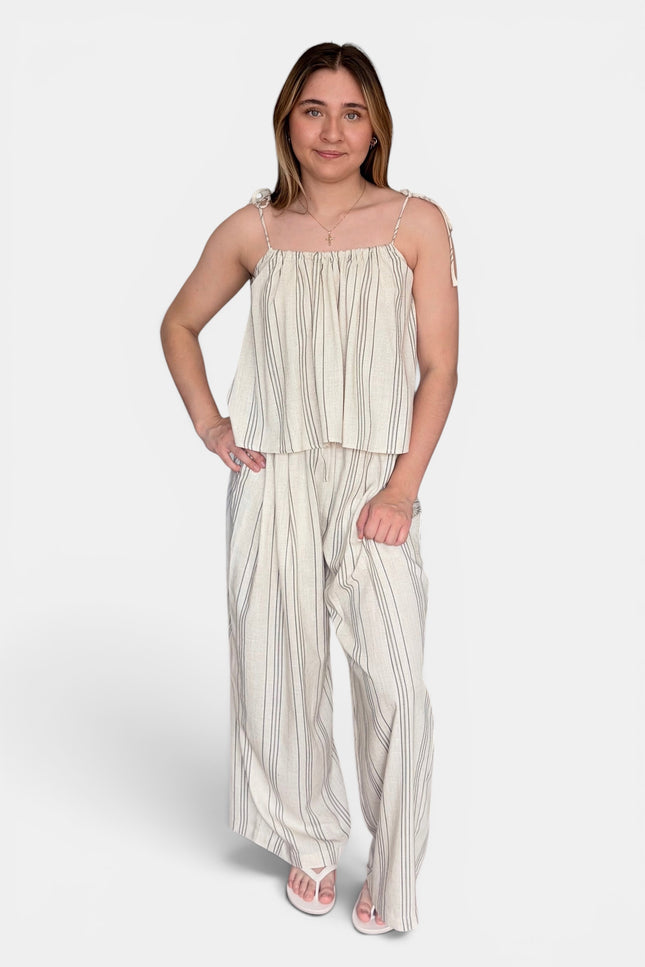 A woman modeling a two-piece matching set consisting of a sleeveless cream and beige vertically striped peplum top and wide-leg trousers, styled with white sandals against a light gray studio background.