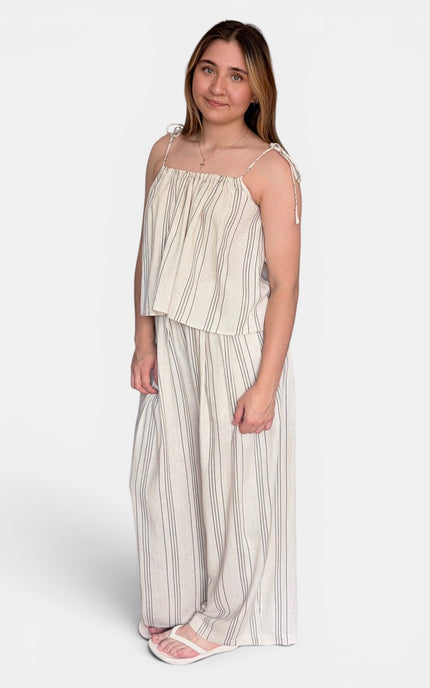 A woman modeling a matching two-piece set consisting of a sleeveless top with thin shoulder ties and wide-leg pants, both in a cream and beige vertical stripe pattern, against a light gray studio background.