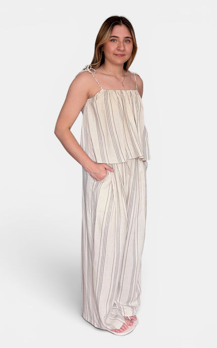 A woman modeling a sleeveless, floor-length maxi dress with thin shoulder straps and vertical stripes in cream and beige, set against a plain light gray studio background.