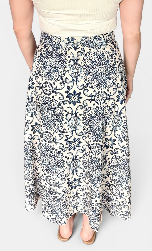 A woman wearing a high-waisted, midi-length A-line skirt with a navy blue and cream floral damask pattern, paired with a cream top and beige heels.