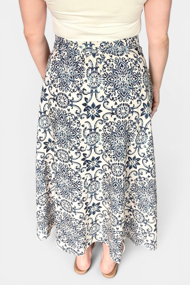 A woman wearing a high-waisted, midi-length A-line skirt with a navy blue and cream floral damask pattern, paired with a cream top and beige heels.