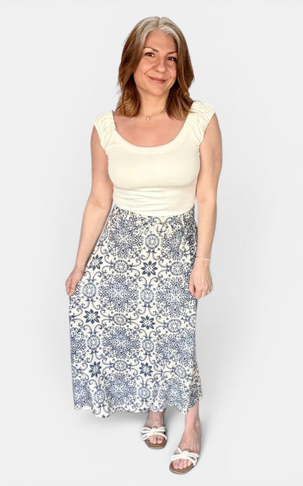 A woman modeling a cream scoop-neck top and a high-waisted midi skirt with a blue and white geometric tile pattern, standing against a light gray background.