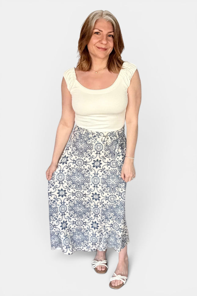 A woman modeling a cream scoop-neck top and a high-waisted midi skirt with a blue and white geometric tile pattern, standing against a light gray background.
