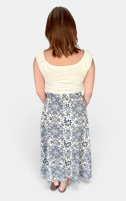 A woman shown from the back wearing a cream scoop-neck top and a white midi skirt with a blue floral and geometric pattern, set against a plain light gray background.