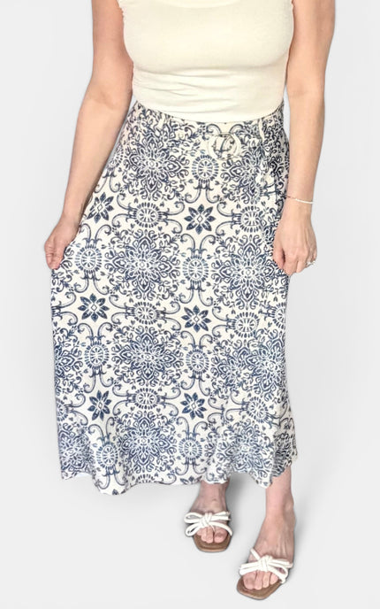 A woman wearing a cream sleeveless top and a long white midi skirt with a blue floral and scroll pattern, paired with white slide sandals, against a light gray background.