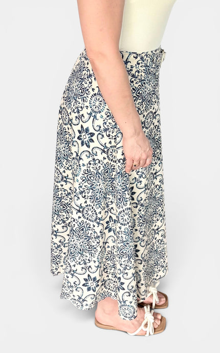 A woman wearing a long, flowing skirt with a navy blue floral and scroll pattern on a cream background, paired with white sandals, shown from the waist down against a light gray studio background.
