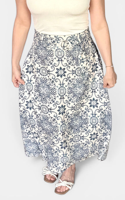A woman wearing a high-waisted, midi-length maxi skirt with a blue and white floral mandala pattern, paired with a cream top and white slide sandals.