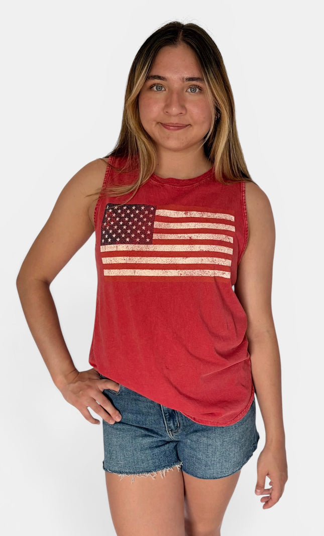 A young woman modeling a red sleeveless muscle tank top with a distressed American flag graphic on the front, paired with denim shorts, against a light gray studio background.