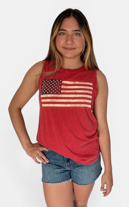 A young woman modeling a red sleeveless muscle tank top with a distressed American flag graphic on the front, paired with denim shorts, against a light gray studio background.