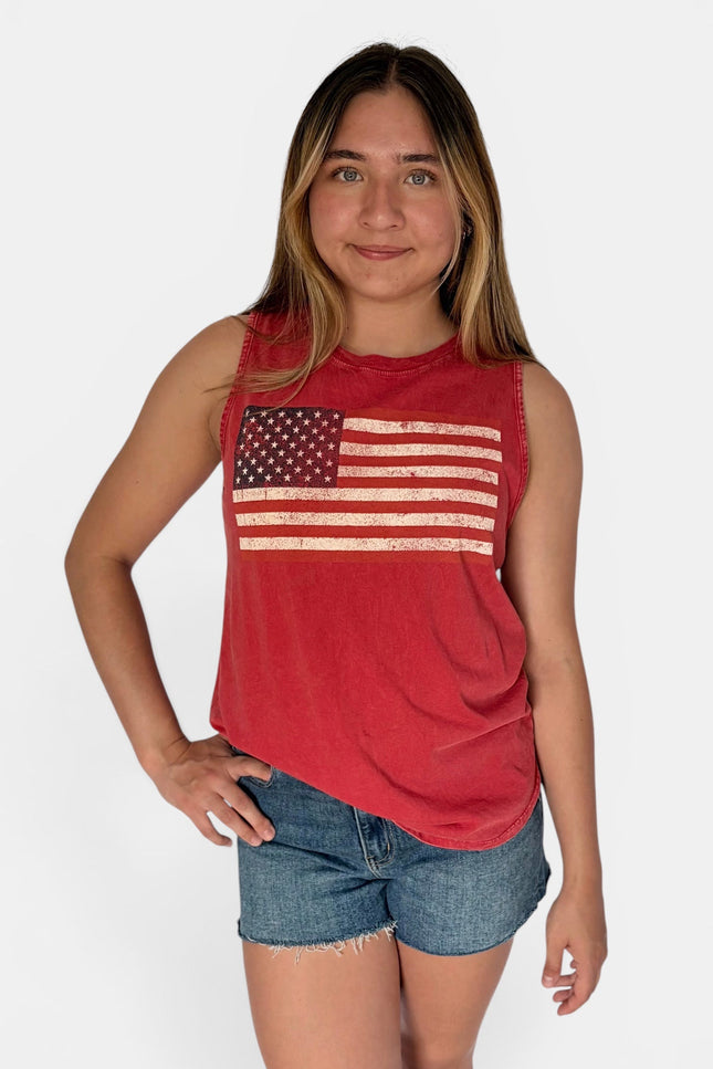 A young woman modeling a red sleeveless muscle tank top with a distressed American flag graphic on the front, paired with denim shorts, against a light gray studio background.