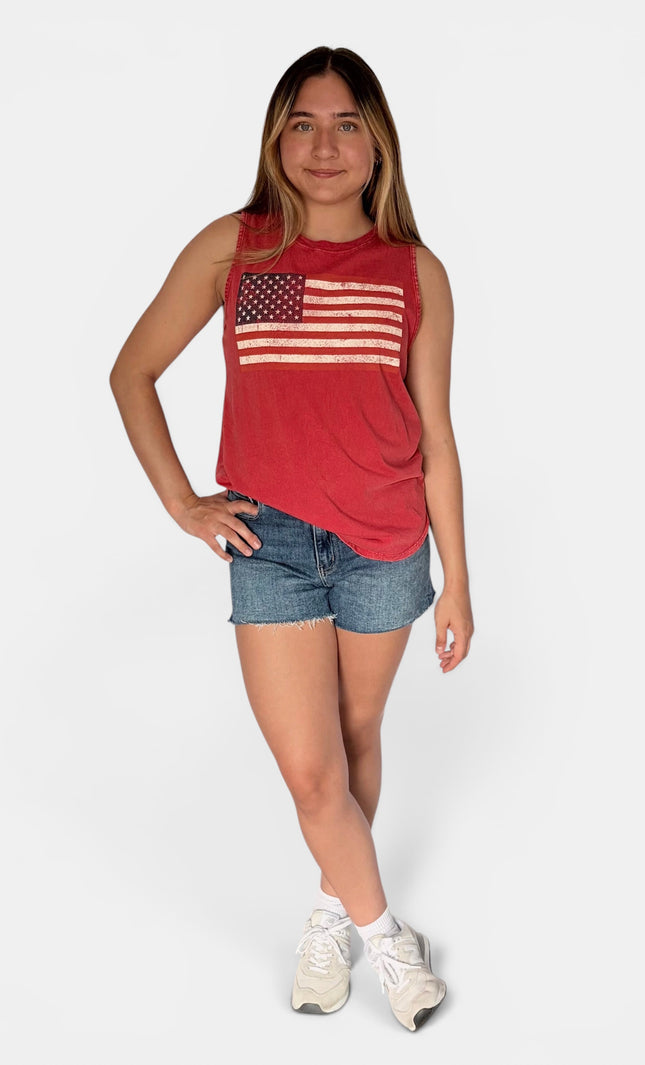 A young woman modeling a red sleeveless tank top with a distressed American flag graphic on the chest, paired with denim shorts and white sneakers, against a light gray studio background.