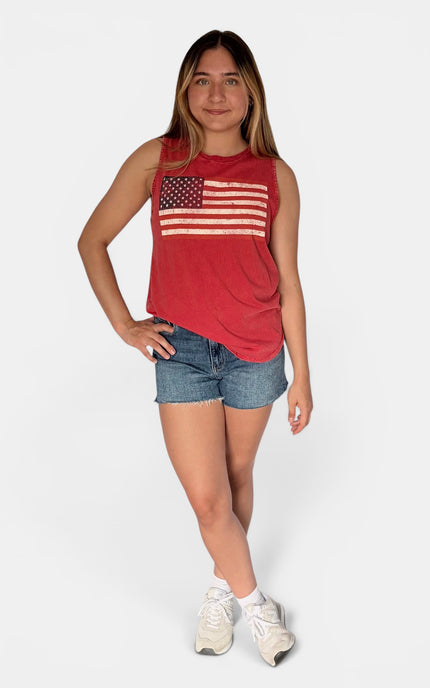 A young woman modeling a red sleeveless tank top with a distressed American flag graphic on the chest, paired with denim shorts and white sneakers, against a light gray studio background.