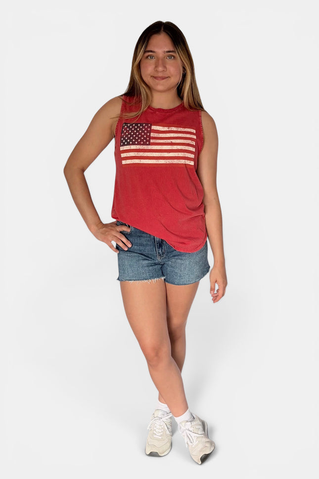A young woman modeling a red sleeveless tank top with a distressed American flag graphic on the chest, paired with denim shorts and white sneakers, against a light gray studio background.
