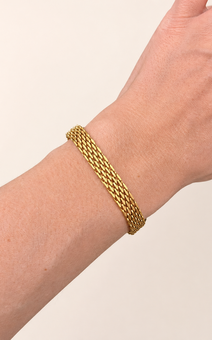 A close-up of a person's wrist wearing an 18K gold plated braided mesh bracelet against a plain light beige background.