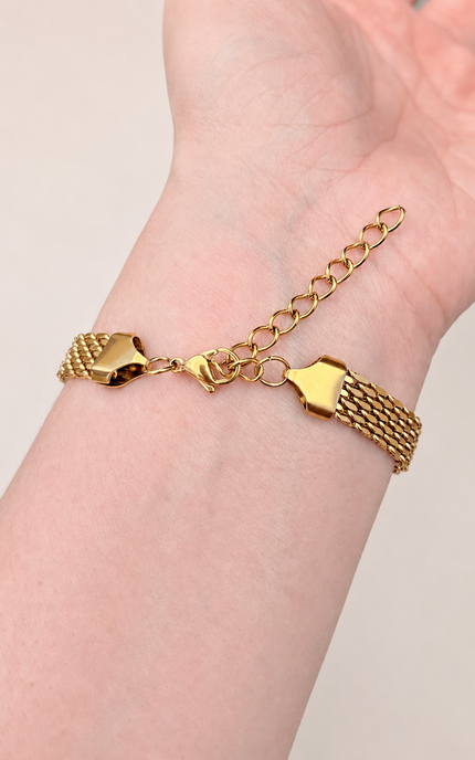 An 18K gold-plated stainless-steel mesh bracelet with a chain extender and polished decorative links, shown worn on a person's wrist against a soft light beige background.