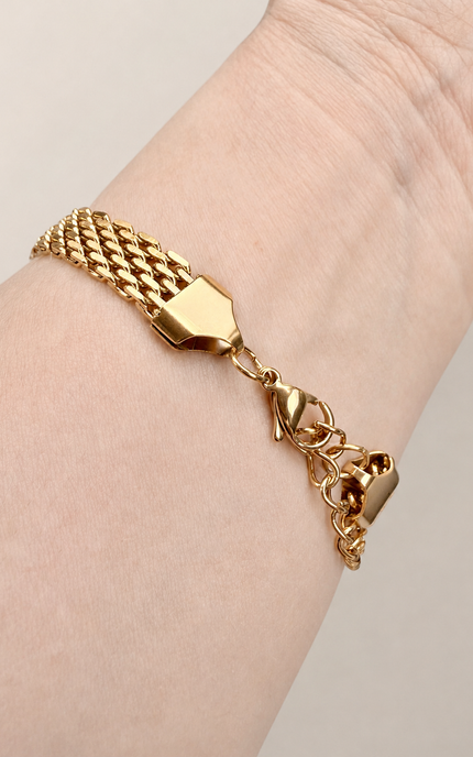 An 18K gold-plated stainless steel chain bracelet with a textured woven section, lobster clasp, and small padlock charm, shown worn on a person's wrist against a light beige background.